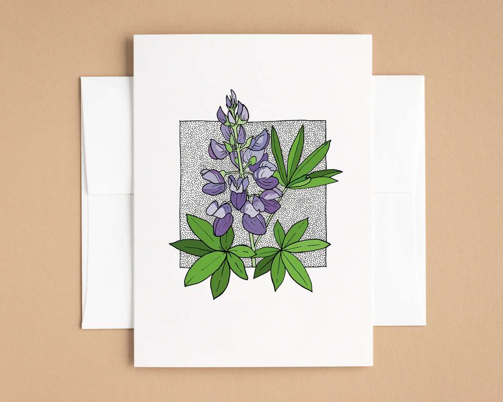 A vertical white card depicts a spray of lupine flowers surrounded by lupine leaves. The card sits on top of a white envelope, which lies on top of a brown backdrop.